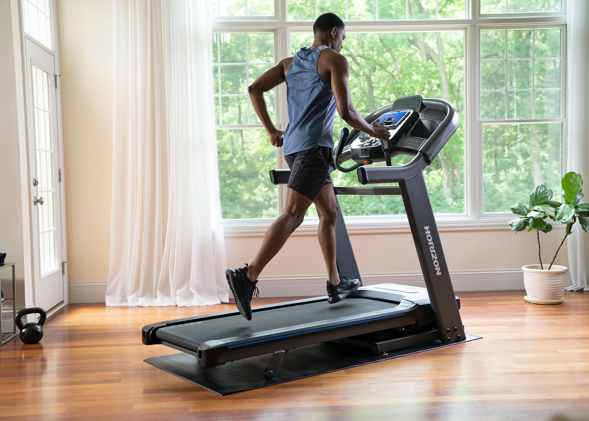 Person using a treadmill in a home setting with large windows.